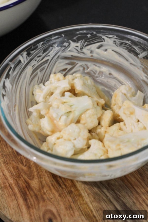 Crispy Bang Bang Cauliflower Oven and Air Fryer 4 Cauliflower pieces being tossed in a glass bowl, coated in a creamy mayonnaise mixture.
