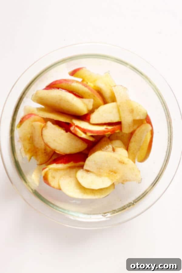Sliced apples mixed with cinnamon and natural sweetener in a clear glass bowl, ready for ramekins.