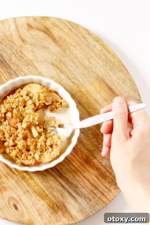 A hand holding a fork, about to scoop a bite of warm, golden brown mini apple crisp from a ramekin.