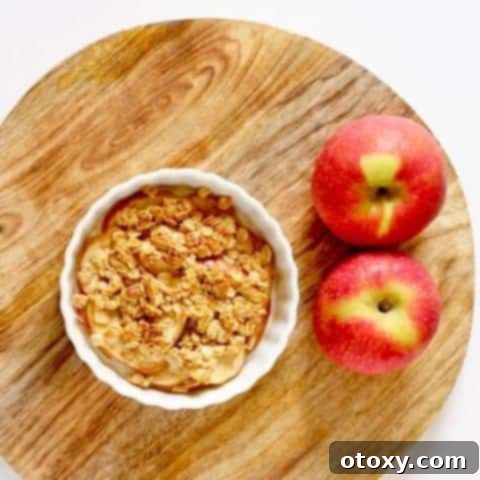 Mini apple crisp in a ramekin on a wooden cutting board, with whole red apples in the background.