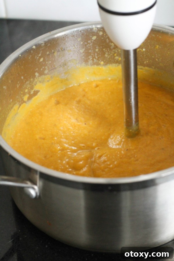 An immersion blender is actively pureeing the cooked pumpkin and cauliflower soup directly in a large pot, creating a smooth, creamy texture.