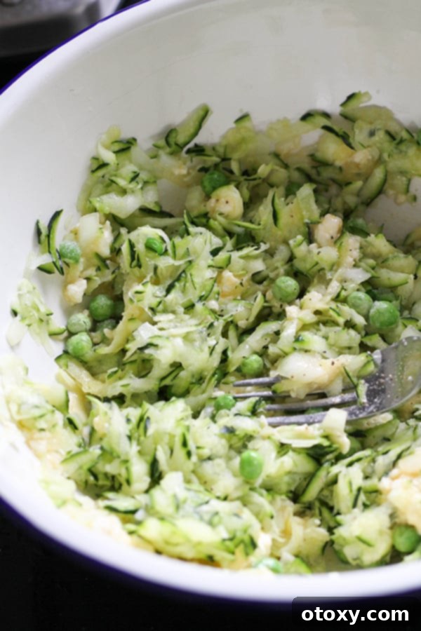 A vibrant mix of fritter ingredients including peas, feta, zucchini, and onion combined in a white mixing bowl, ready for flour.