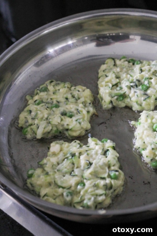 Multiple pea & feta fritters sizzling in a stainless steel frying pan, turning golden brown as they cook.