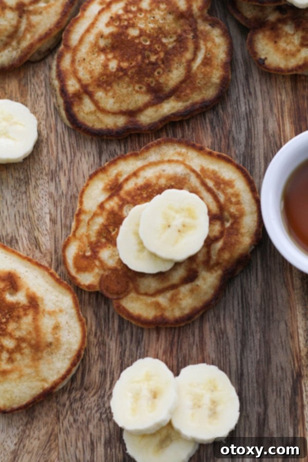 Freshly cooked banana pikelets generously topped with sliced bananas and a drizzle of maple syrup, artfully arranged on a rustic wooden board.