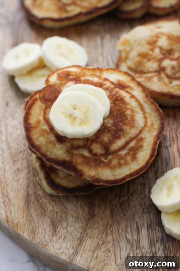 A beautifully stacked tower of golden banana pikelets, garnished with fresh banana slices and a delicate drizzle of honey, resting on a textured wooden board.