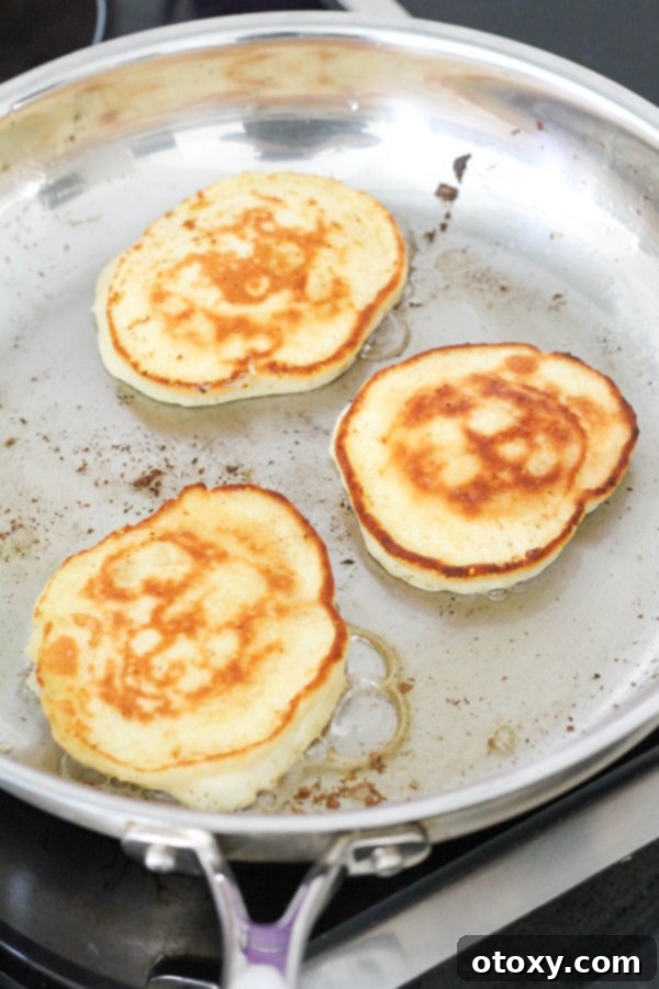 Three perfectly golden banana pikelets sizzling in a hot frying pan, with one already flipped, displaying a beautifully browned surface, ready to be transferred.