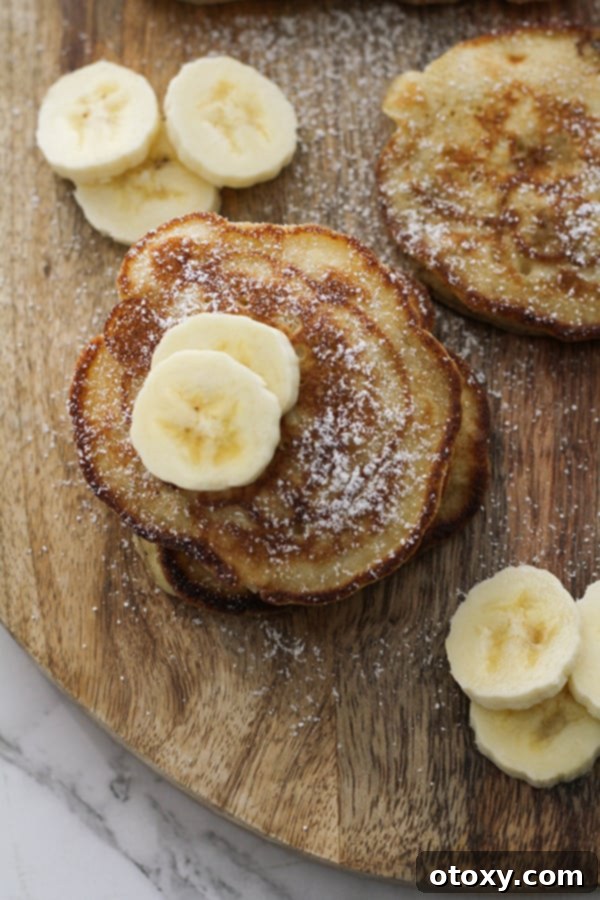 A neat stack of golden-brown banana pikelets, elegantly drizzled with maple syrup and garnished with fresh banana slices, presented on a wooden cutting board.