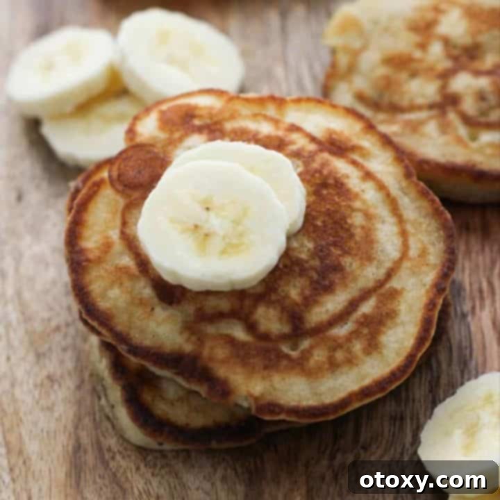 A stack of banana pikelets, glistening with syrup, presented on a wooden board with fresh banana slices beside it.