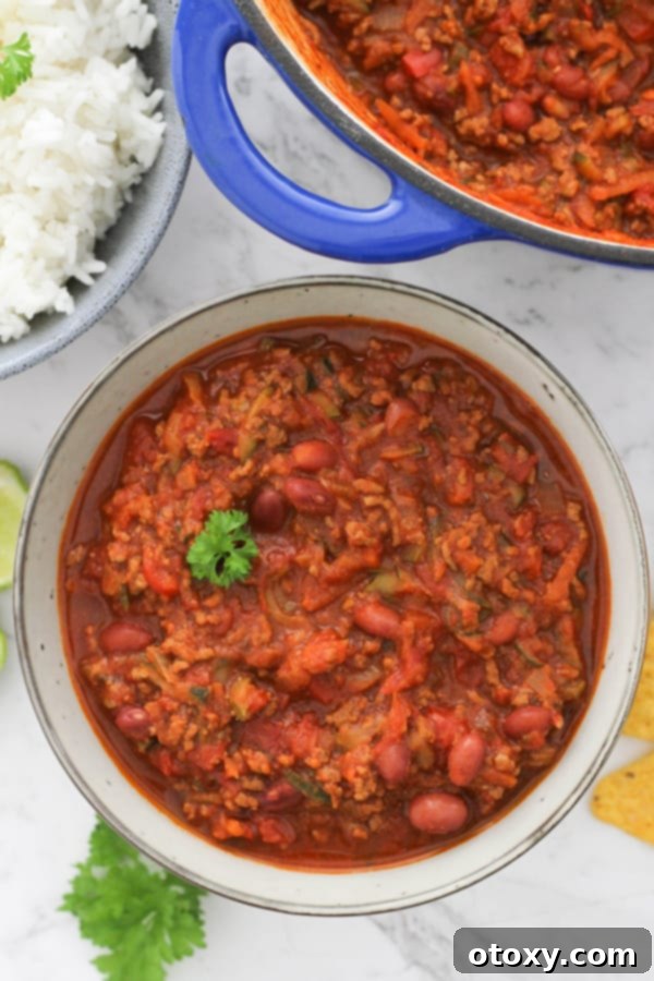 A vibrant bowl of healthy Chilli Con Carne, garnished with fresh herbs, beside a bowl of fluffy rice and a large pot of the rich stew on a countertop.