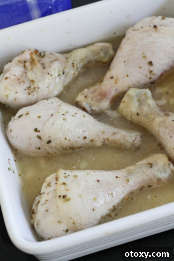 Chicken drumsticks are placed on top of uncooked rice and chicken stock in a baking dish, ready to return to the oven.