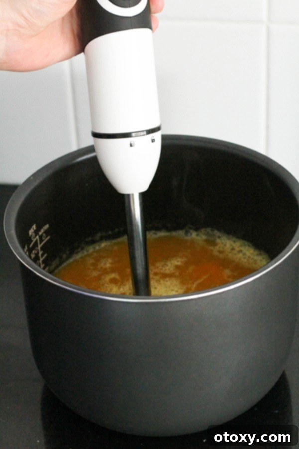 A hand holding an immersion blender, actively blending the cooked pumpkin soup directly in the pot to achieve a smooth texture.