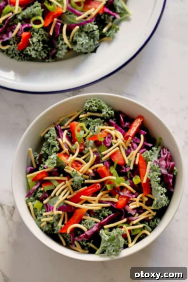 A beautifully arranged crunchy kale salad with lemon tamari dressing in a white bowl, garnished with fresh ingredients. A side plate with more salad is visible in the background.