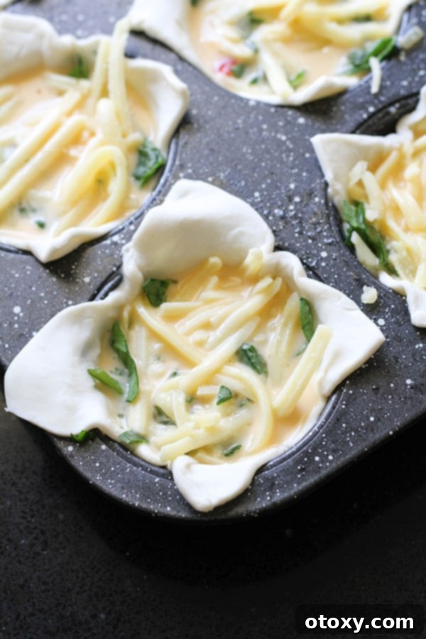 Mini quiches in a muffin tray, with puff pastry, vegetables, cheese, and egg custard poured in, ready for baking.