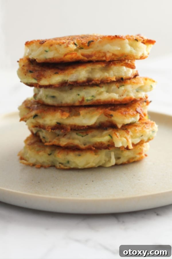 A perfectly stacked pile of golden-brown potato and zucchini fritters on a pristine white plate, garnished with fresh herbs, ready to be served.