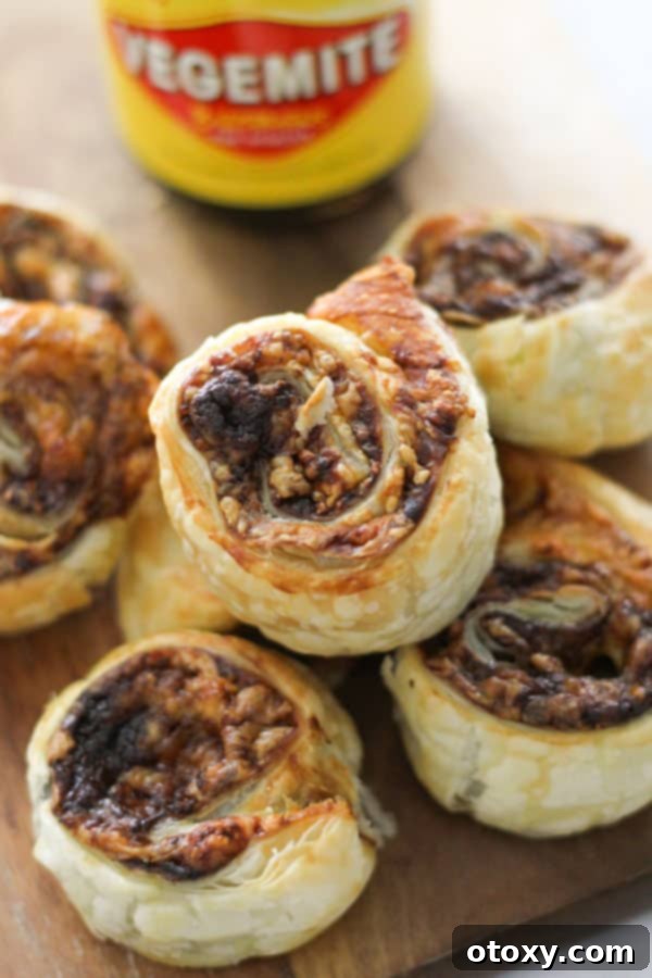 Easy Vegemite and Cheese Pinwheels Baked or Air Fried 2 Freshly baked Vegemite and cheese pinwheels stacked on a rustic wooden board, showcasing their golden crust and swirled layers.