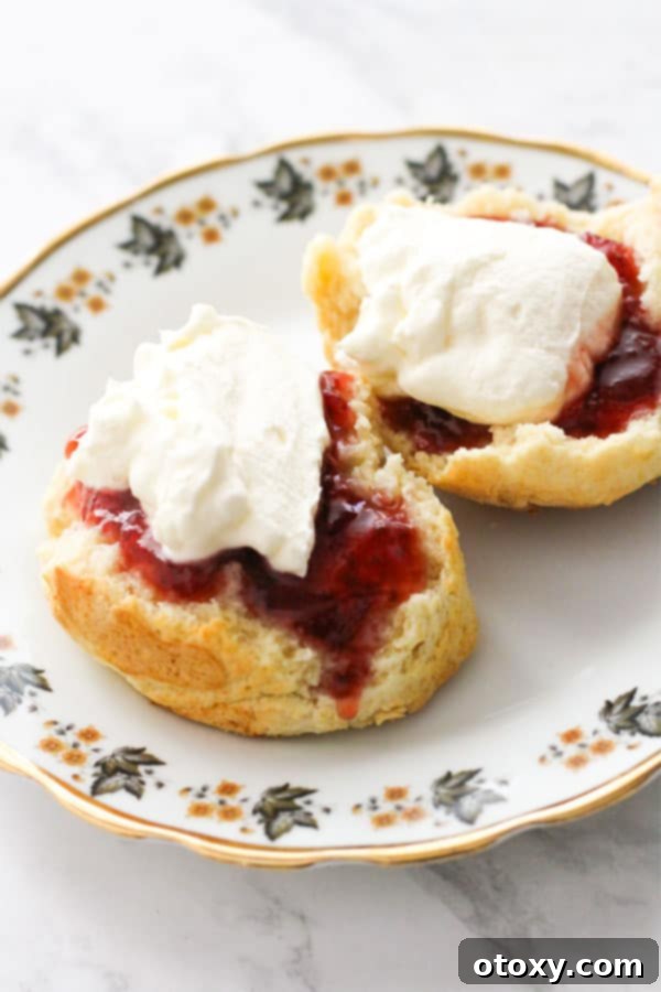 Golden Air Fryer Scones 10 A close-up shot of a perfectly baked scone, artfully adorned with a dollop of fresh cream and a spoonful of glistening jam, resting on a white plate, invitingly presented.