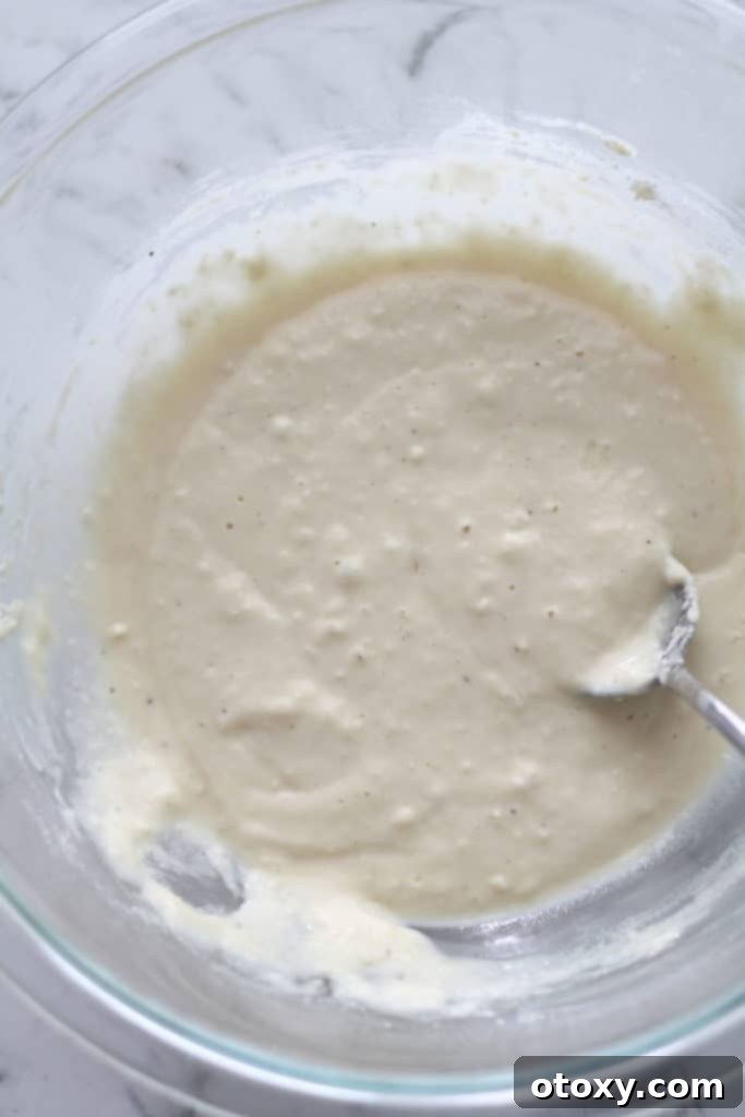 A close-up of thick, creamy fritter batter in a clear glass bowl, showing its consistency.