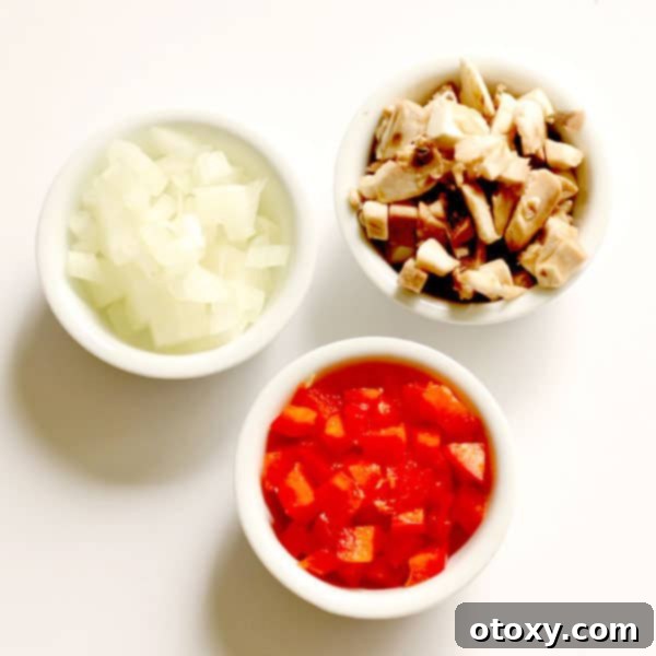 Three small white bowls filled with diced mushrooms, onions, and red capsicum (bell pepper) on a white tabletop, prepared for cooking.