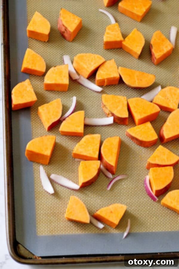Diced sweet potatoes and thinly sliced red onion seasoned with olive oil on a baking tray lined with parchment paper, ready for roasting.