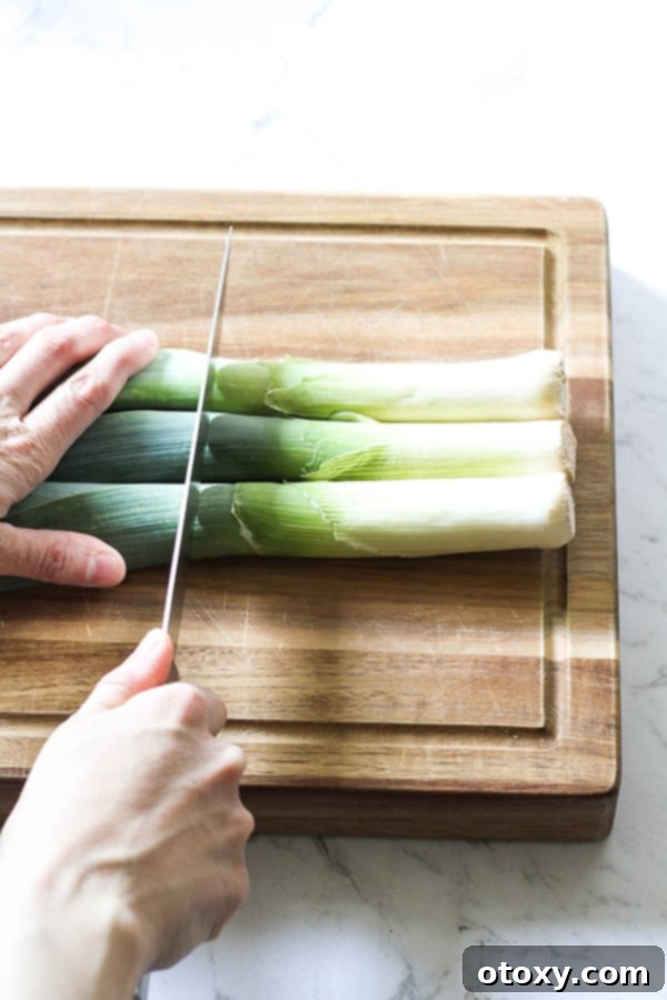 Leeks on a wooden chopping board with a hand precisely cutting off the tough dark green ends.