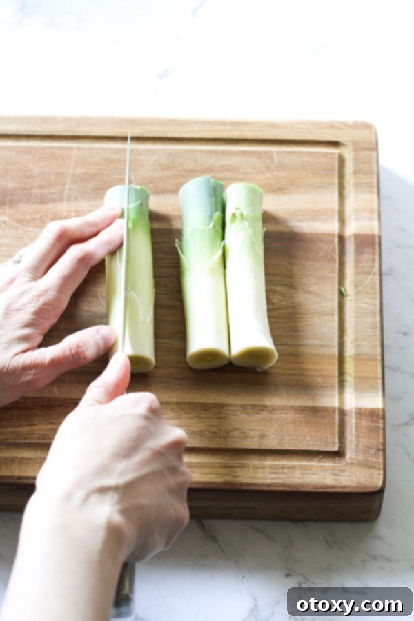 Leeks on a wooden chopping board with a hand expertly slicing them in half lengthwise.