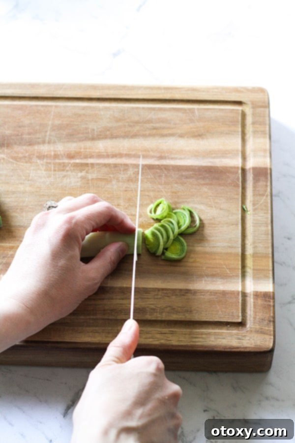 Leeks on a wooden chopping board with a hand uniformly slicing the leeks into delicate half moons.