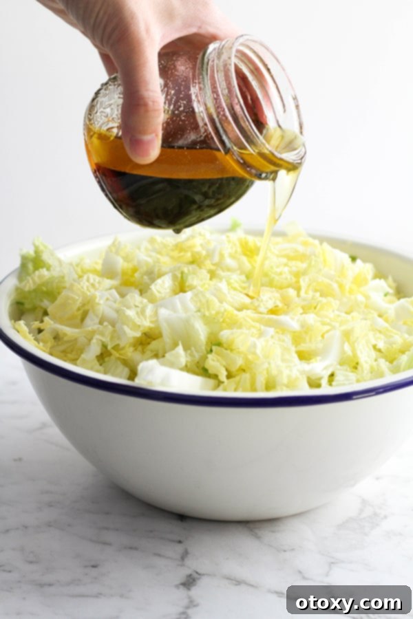 A hand pouring sesame soy dressing over a bowl of cabbage and crunchy noodle salad ingredients.