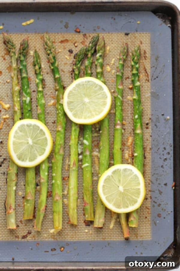 asparagus spears laid out in a single layer on a baking tray covered in parmesan cheese, minced garlic and lemon slices