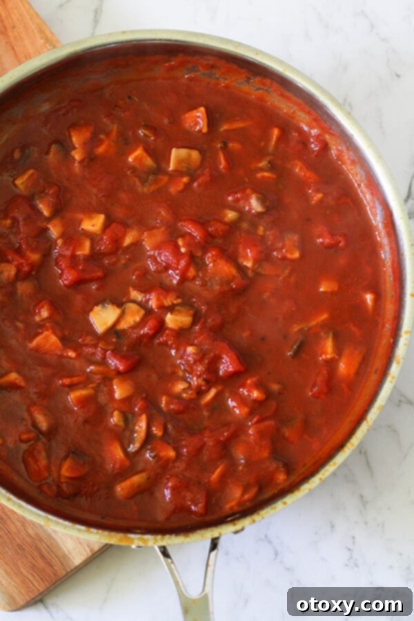 A vibrant tomato-based sauce for bacon and mushroom pasta simmering in a large, dark saucepan, ready to be combined with pasta.