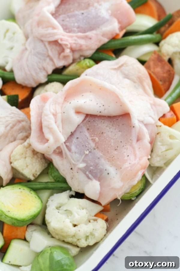Sheet Pan Golden Chicken Thighs with Roasted Vegetables 7 A chicken thigh on a baking tray, skin side up with the skin peeled back to expose the meat, being patted dry with a paper towel. Vegetables are visible underneath.