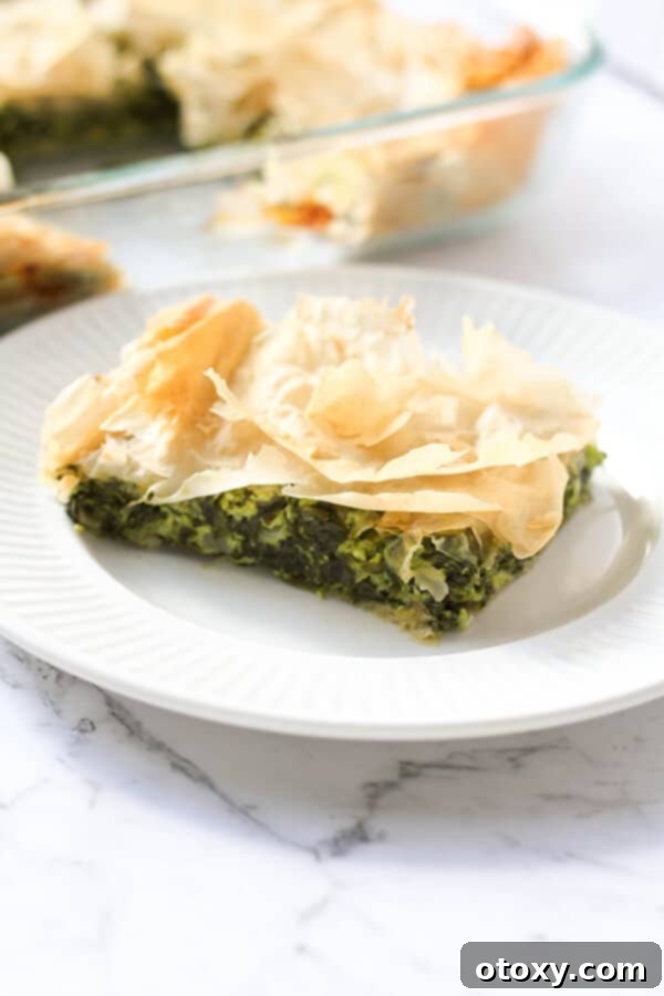 A perfectly baked slice of Spanakopita on a white plate, with a full tray of the golden pie in the background, showcasing its flaky texture.