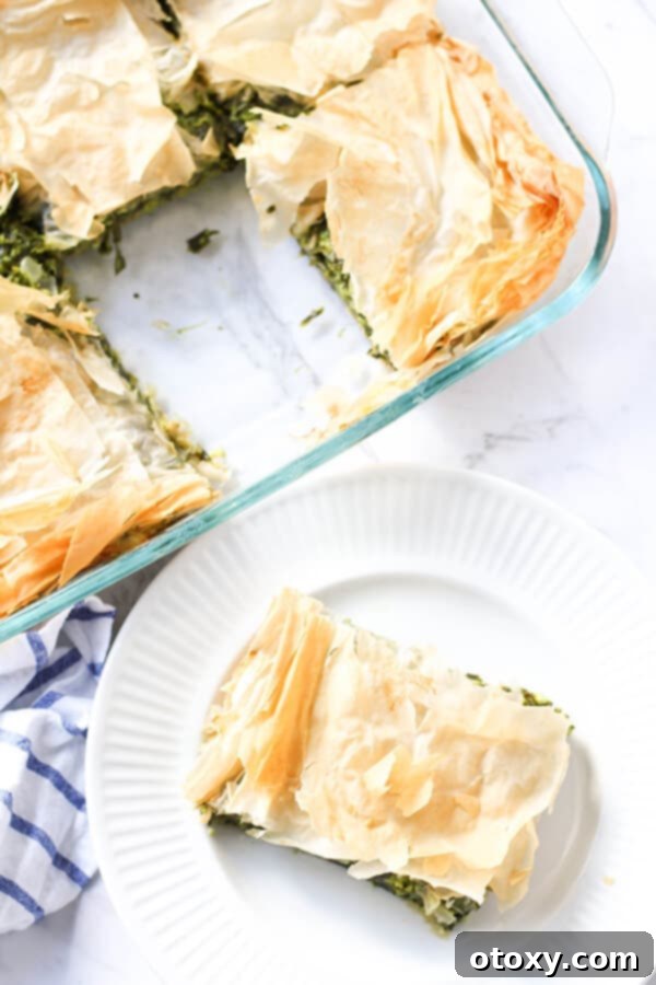 A close-up shot of a Spanakopita slice, revealing the layers of golden filo pastry and the vibrant spinach and cheese filling, resting on a white plate with more pie in the background.