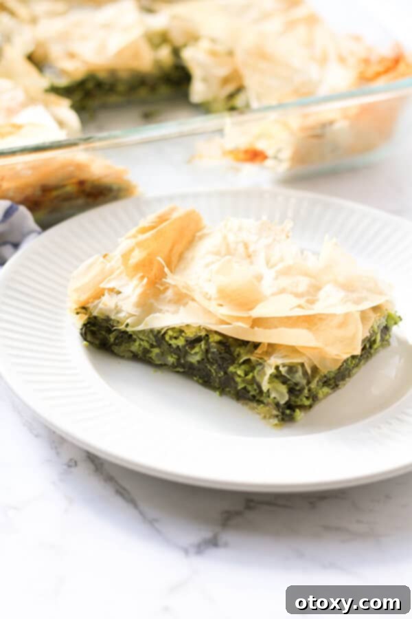 A delectable slice of golden Spanakopita resting on a pristine white plate, with the full, baked pie tray blurred in the background, highlighting the pie's flaky texture.
