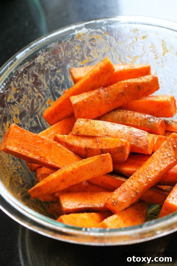 Sweet potato wedges in a glass mixing bowl, thoroughly coated with olive oil and a blend of spices.