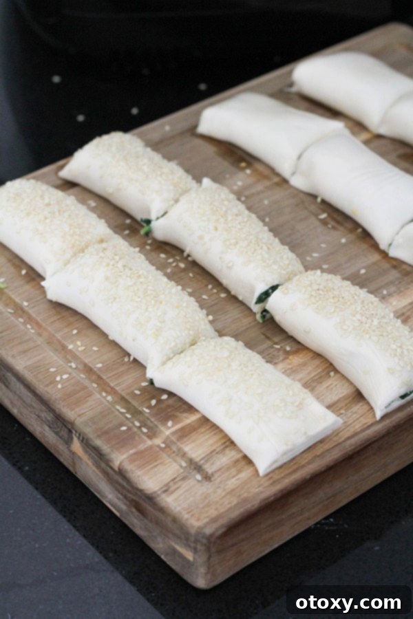Spinach Ricotta Rolls Oven or Air Fried 8 Uncooked spinach and ricotta rolls neatly arranged on a cutting board, brushed with egg wash and topped with sesame seeds, ready for baking.