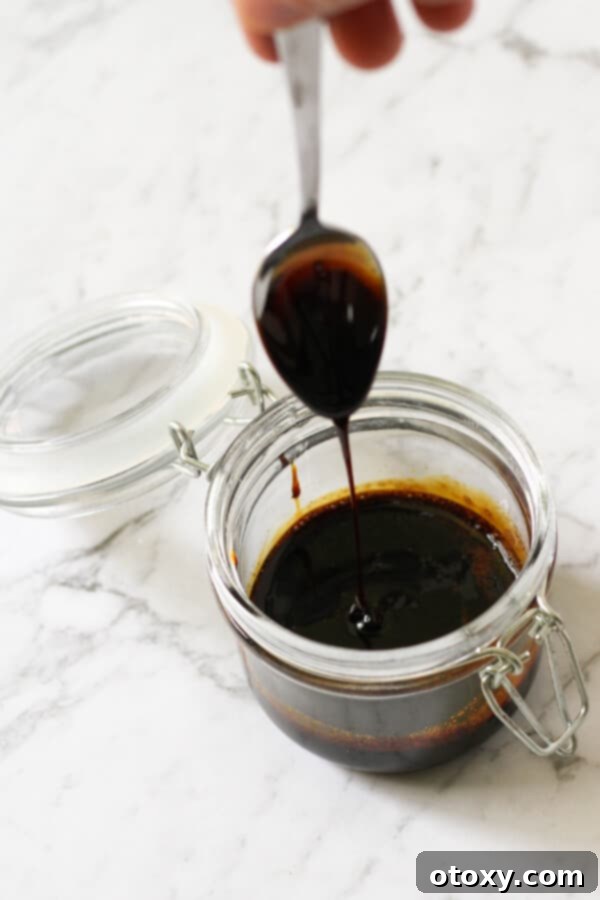 Close-up of balsamic glaze in a clear glass jar, with a spoon lifted slightly, showing its thick, glossy consistency.