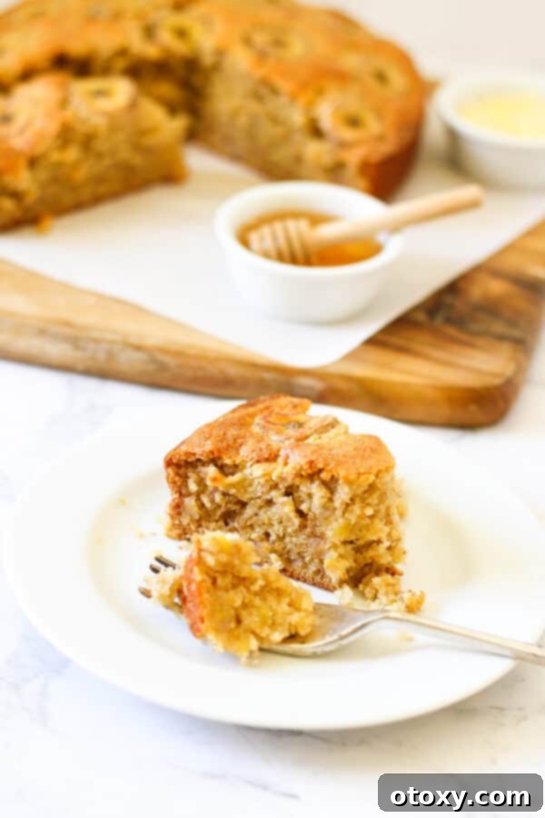 a slice of banana cake on a white plate with the rest of the cake on a wooden board behind.
