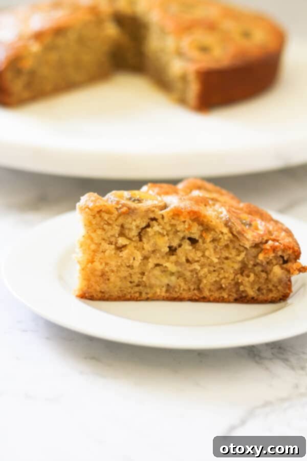 a slice of banana cake on a white plate with the rest of the cake on a wooden board behind.
