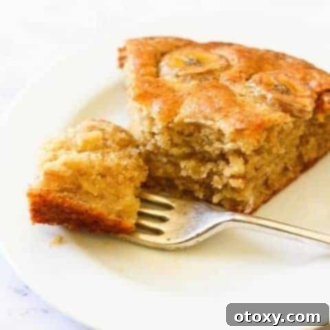 A beautiful slice of moist banana cake with caramelized banana topping on a white plate, with the rest of the cake blurred in the background on a wooden board.