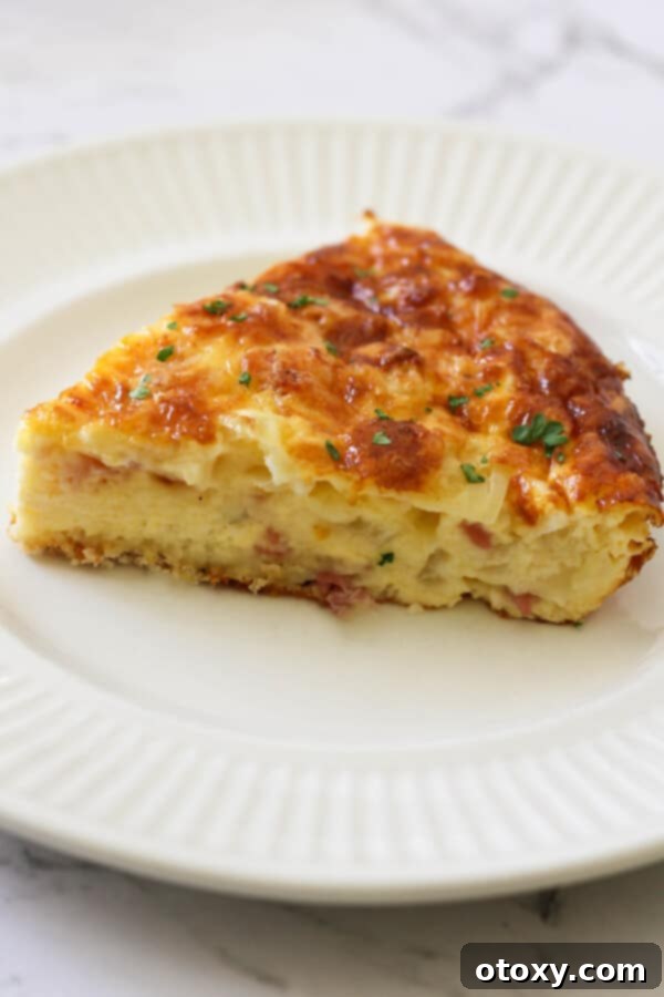 A close-up of a single slice of Impossible Quiche on a white plate, revealing its fluffy interior, savory ham, and melted cheese fillings.