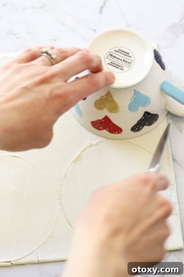 A hand using a round cookie cutter to cut circles from a sheet of puff pastry.