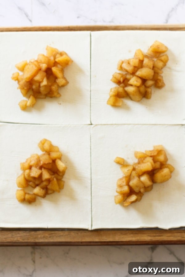 Puff pastry squares prepared on a surface, each with a spoonful of diced cinnamon apple filling in the center, ready for folding.
