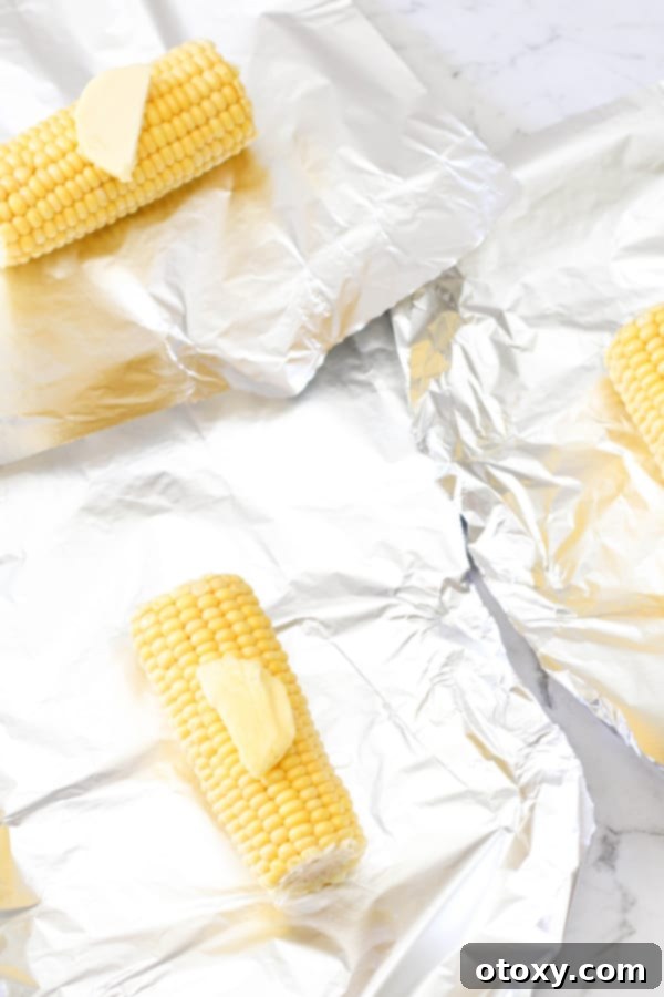 Shucked corn on the cob placed on a sheet of aluminum foil with a pat of butter, ready for wrapping.