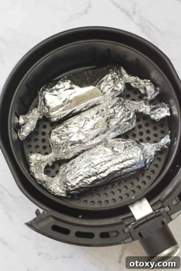 Three foil-wrapped corn on the cobs arranged inside an air fryer basket during cooking.