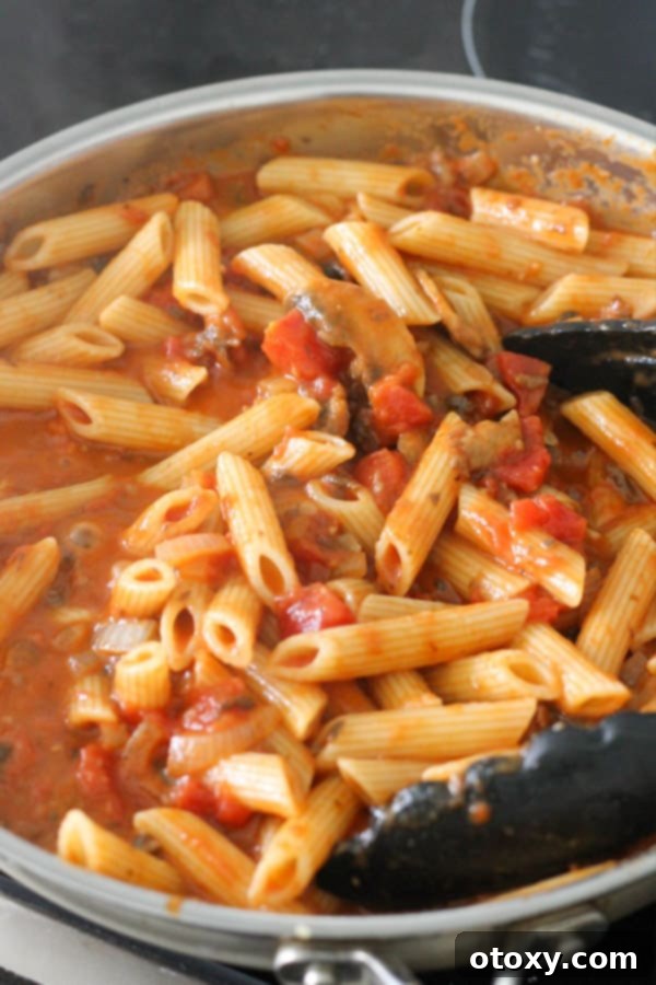 Al dente penne pasta combined with the creamy tomato mushroom sauce in a large frying pan, being tossed to ensure every strand is perfectly coated.