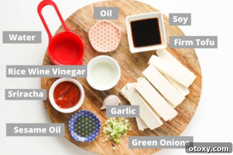 A rustic wooden board showcasing all the fresh ingredients needed to make spicy tofu: olive oil, firm tofu, sriracha, soy sauce, rice wine vinegar, sesame oil, green onions, and garlic.